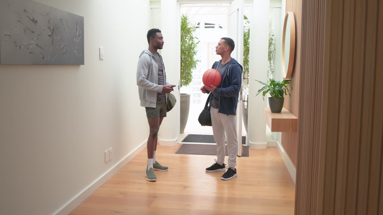 Amici maschi di diverse etnie che tornano a casa, aprendo la porta con un pallone da basket e borsoni, parlando e camminando.