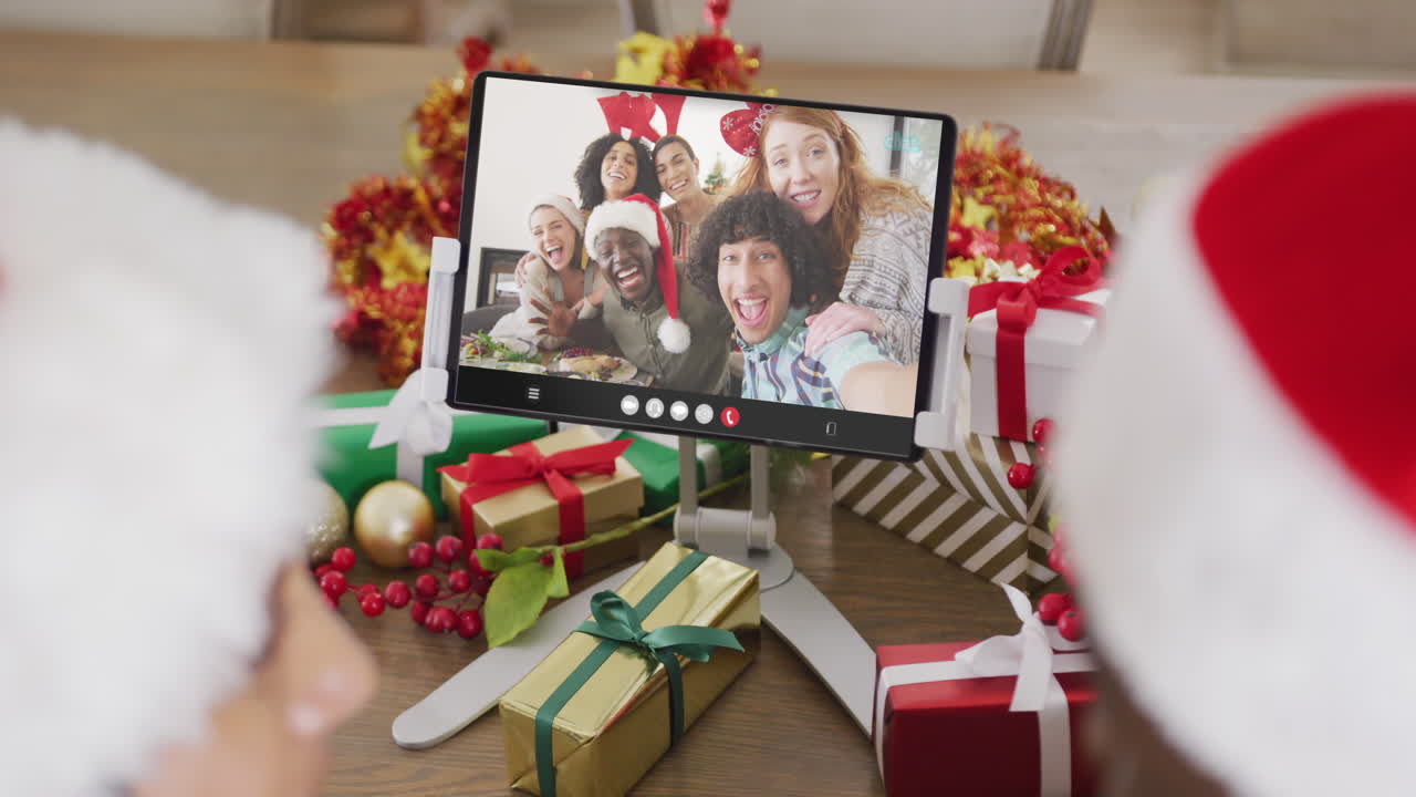 una pareja caucásica con sombreros de santa haciendo una llamada de video de tableta con amigos felices y diversos