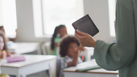 Video-of-hands-of-caucasian-female-teacher-holding-tablet-in-class-of-diverse-pupils