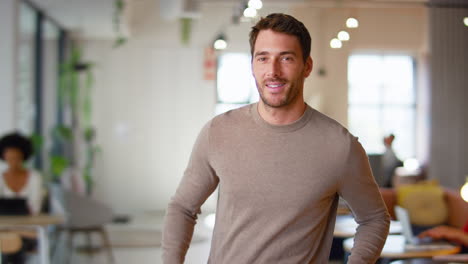 Portrait-Of-Smiling-Businessman-Standing-In-Busy-Modern-Open-Plan-Office