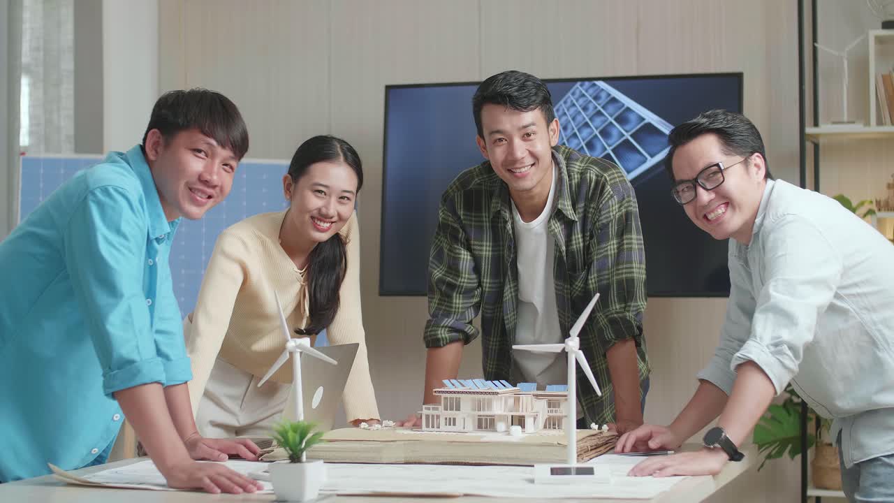 아시아 엔지니어 그룹은 사무실에서 태양 전지판을 가진 작은 집 모델에 대해 일하면서 카메라에 미소 짓고 있습니다.