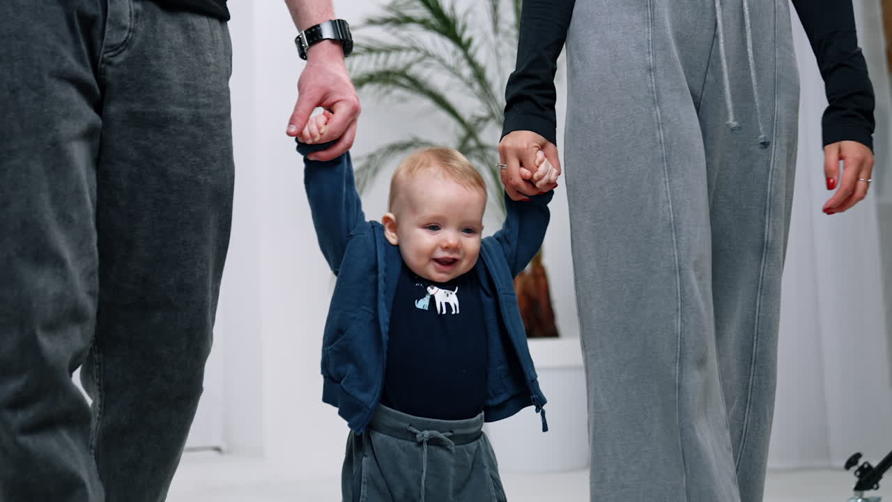 Unrecognized parents lead their cute little baby by hands. Smiling kid is happy to make first steps.