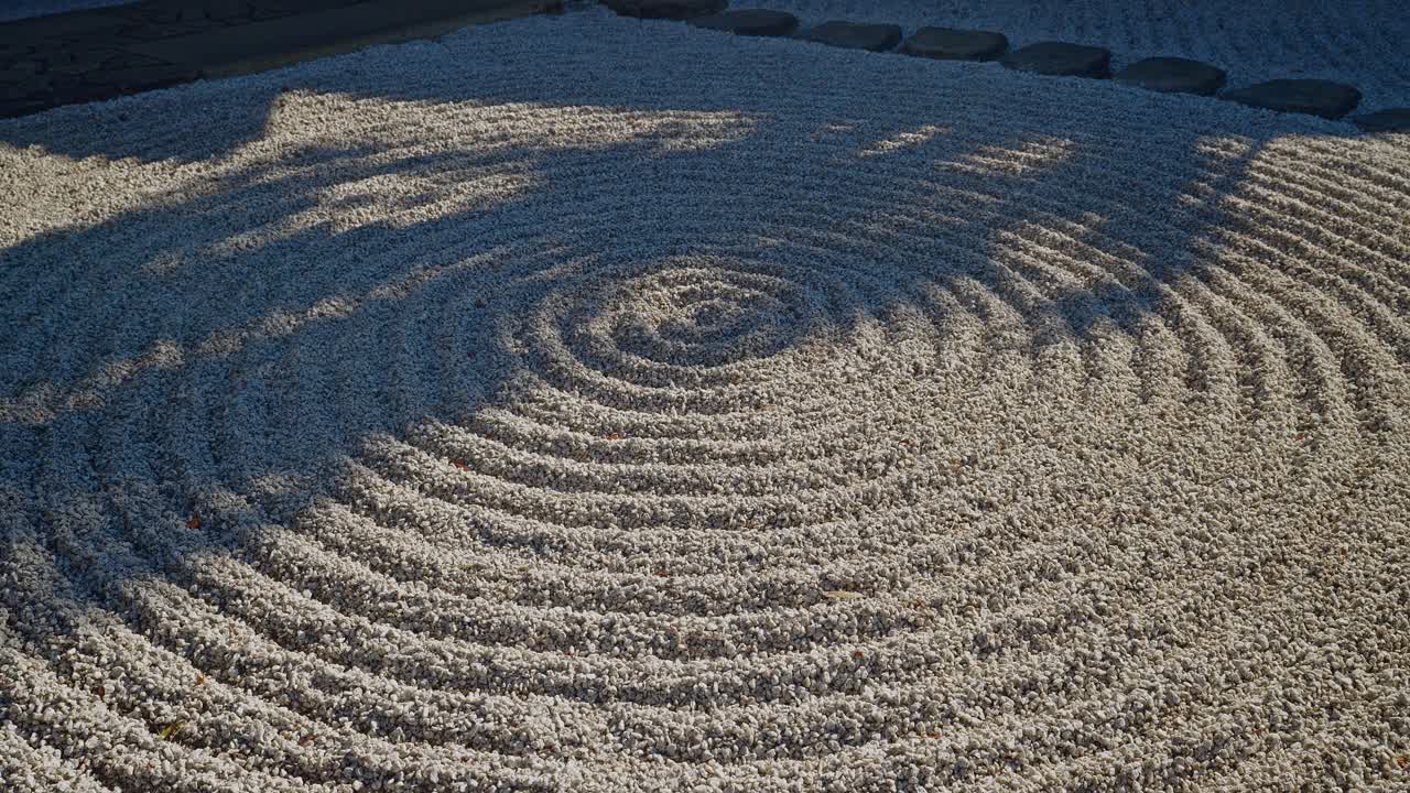 Eine detaillierte Nahaufnahme eines Karesansui-Trockengartens. Sonnenlicht hebt die komplizierten kreisförmigen geharkten Kiesmuster und Wellen im weißen Sand hervor und schafft eine meditative und friedliche Atmosphäre.