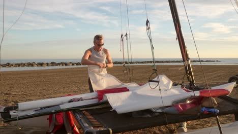 man gathering the sails of a catamaran on the beach at sunset