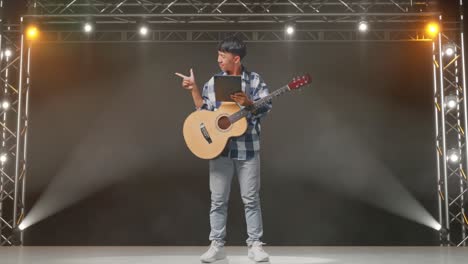cuerpo lleno de adolescente asiático con guitarra sosteniendo una tableta y apuntando a un lado mientras está de pie en el escenario