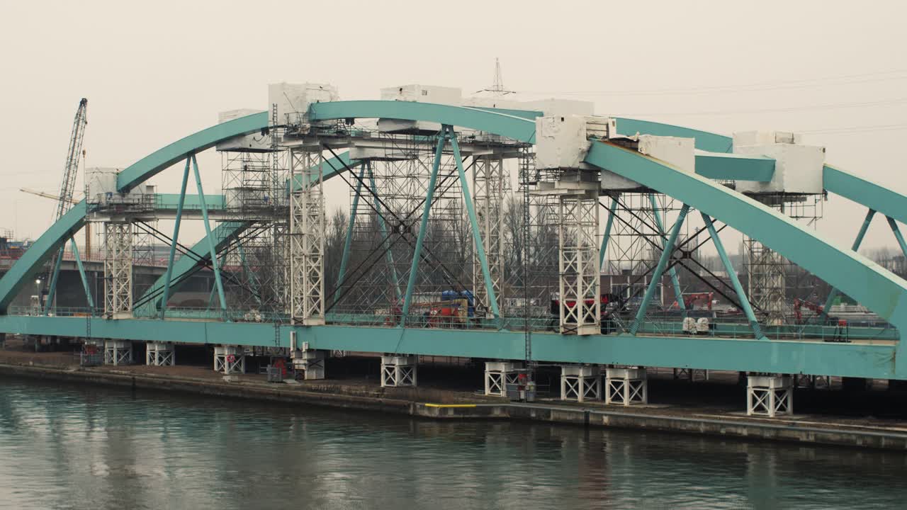 Static shot of a large bridge frame with aqua steel beams, scaffolding and construction equipment over a canal