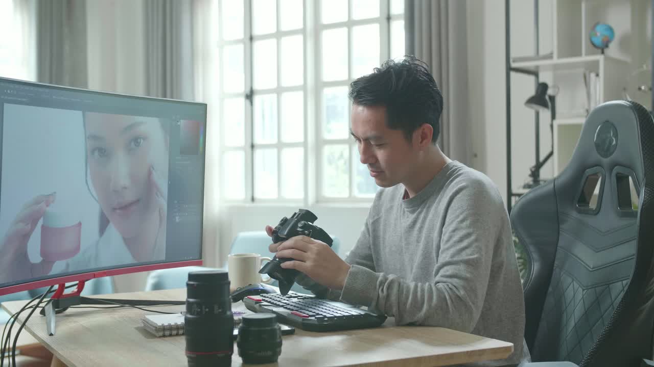 Asian Cameraman In Long Sleeved T-Shirt Holding And Looking At Camera While Using Desktop Computer For Editing Photo At Home.