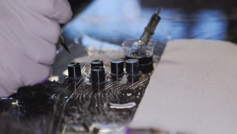 Tattoo artist gloved hand dipping single needle tip in one of many black ink cups on sterilized cling wrapped shop studio space