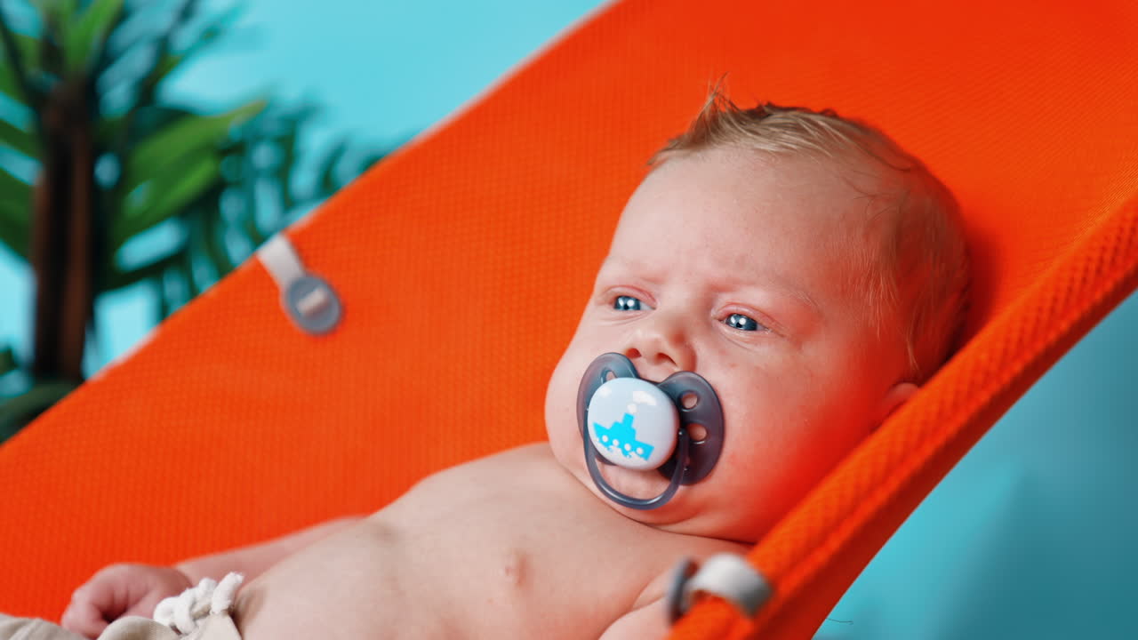 Unhappy blue-eyed blond Caucasian infant with a pacifier in mouth. Portrait of a kid in orange chair. Close up.