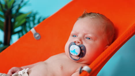unhappy blue-eyed blond caucasian infant with a pacifier in mouth. portrait of a kid in orange chair. close up.