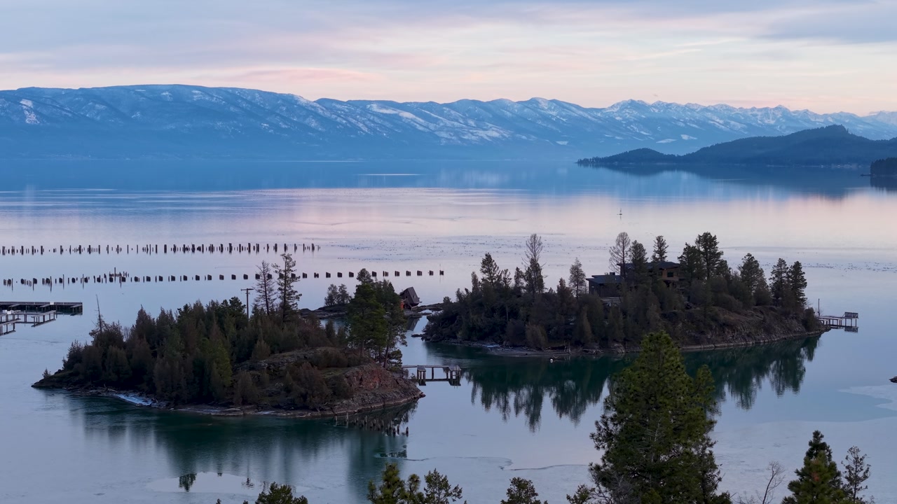 Drone footage gliding above a tranquil lake scattered with small tree-covered islands and rows of wooden posts, with snowy Rocky Mountains in the distance.