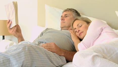 Man-reading-a-book-in-bed-while-his-wife-sleeps-on-his-shoulder