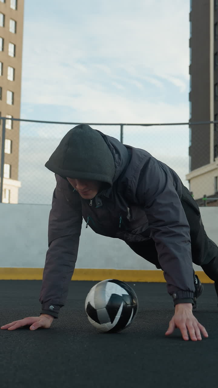 Sportsman performing dynamic push-ups alternating hand placement on soccer ball, showcasing strength, coordination, and agility, background includes goal post and residential buildings