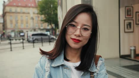 검은 머리에 안경을 쓰고 카메라에 포즈를 취하고 도시에서 야외에서 웃고 있는 매력적인 젊은 여성의 초상화