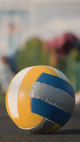 close-up of volleyball on outdoor court with blurred view of athletes bending during workout in background, building and sports equipment visible, vibrant sunlight illuminating scene