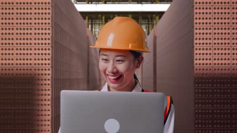 close up of asian female engineer with safety helmet working on a laptop while standing with red brick packed in stacks are stored
