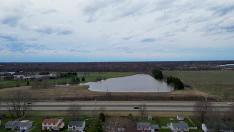 An-aerial-footage-showcasing-a-residential-neighborhood-adjacent-to-a-highway,-with-a-water-body-and-open-fields-in-the-background