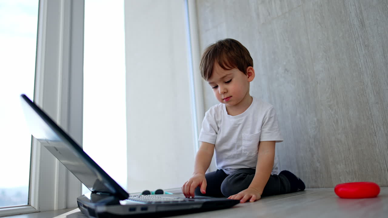 Adorable baby boy sitting at the laptop on the floor. Calm kid presses keys, touchpad and closes computer. Low angle view.