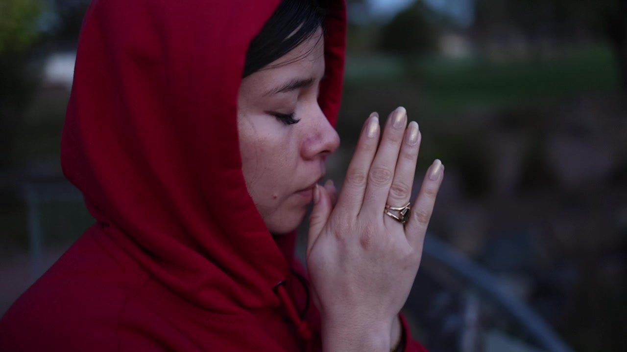 Seorang wanita berjaket hoodie merah berdiri merenung di jembatan kayu dekat air saat senja, mata terpejam dalam doa. Cahaya senja alami yang lembut, berlatar taman luar ruangan, menangkap suasana hati yang reflektif.