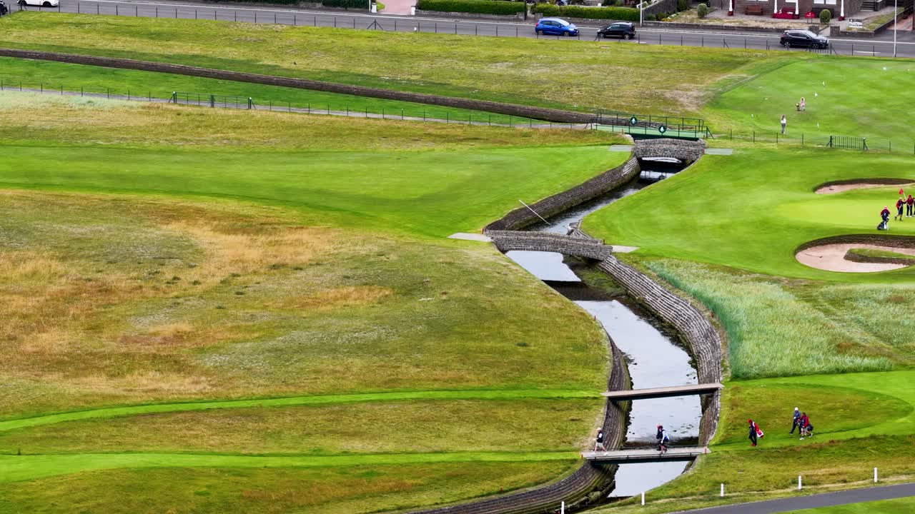 Golfers and caddies walk across stone bridge on lush fairway, aerial drone view, daylight
