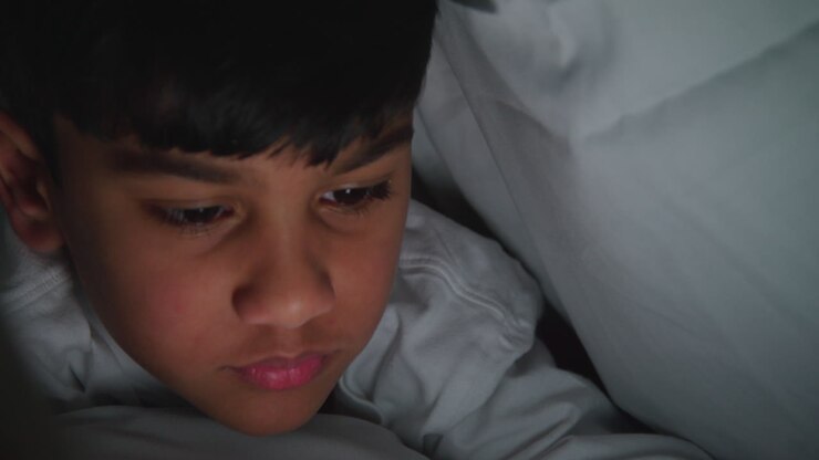 Primo piano di un giovane ragazzo nella sua camera da letto a casa che usa il telefono cellulare per inviare messaggi sotto le coperte o il piumone di notte 5