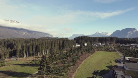 Aerial-drone-of-beautiful-alpine-mountain-range-in-the-distance-with-grassy-fields,-trees-and-a-town