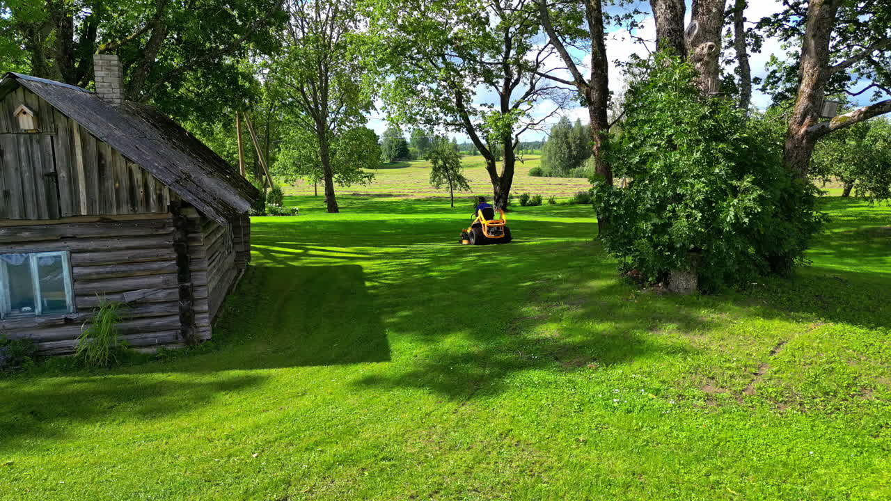 Man mowing grass with a riding lawnmower beside a small wooden house on a sunny day