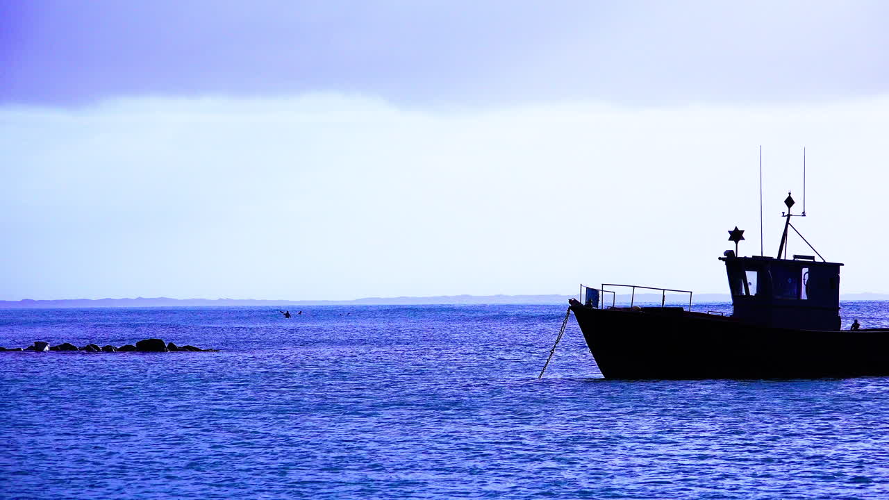 A fishing boat drifts on anchor in the evening in a calm bay with surfers in the distance