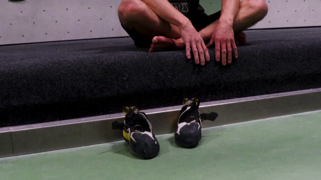 Video di una persona seduta sul pavimento di una palestra di bouldering con le scarpe davanti.