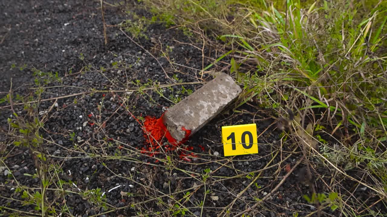 Top down view of a yellow numbered police investigation crime scene marker placed next to a bloody brick or blunt force object murder weapon outdoors on dark gravel and grassy ground at day time