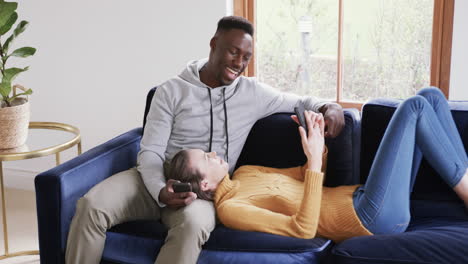 Happy-diverse-couple-sitting-and-lying-on-sofa-and-using-smartphones-in-home,copy-space