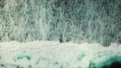 White-foam-of-ocean-waves-splashing-on-shallow-turquoise-lagoon-with-rocky-seabed,-sea-texture-seen-from-above-in-Caribbean