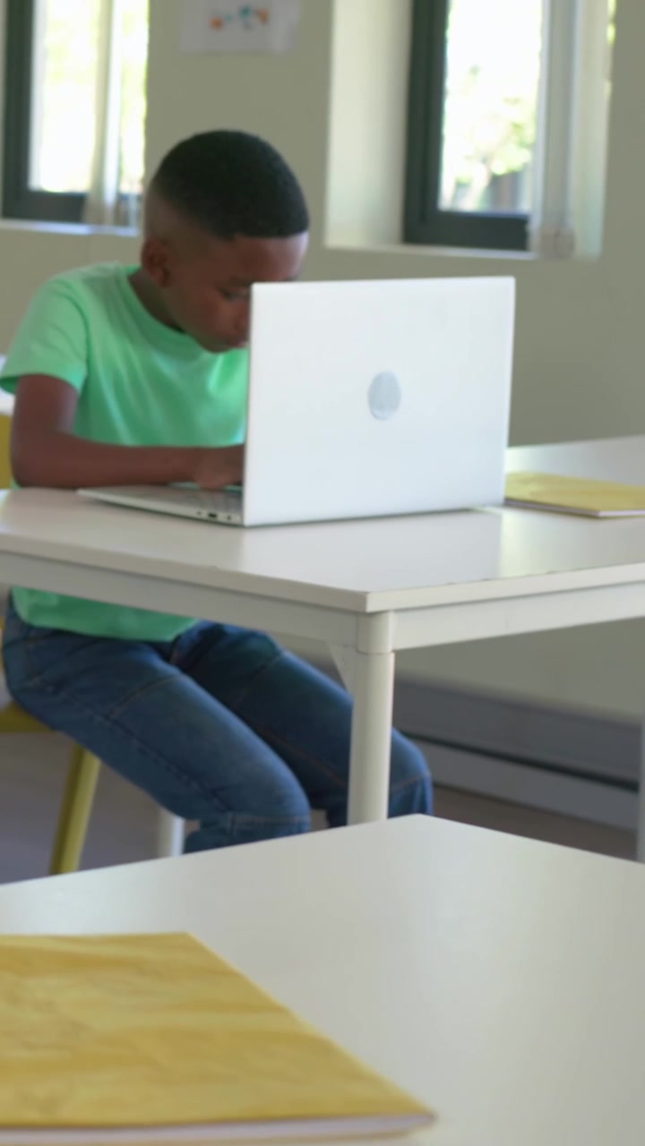 Video verticale: La telecamera inquadra un adolescente con una maglietta verde che digita su un laptop bianco in classe mentre fa i compiti.