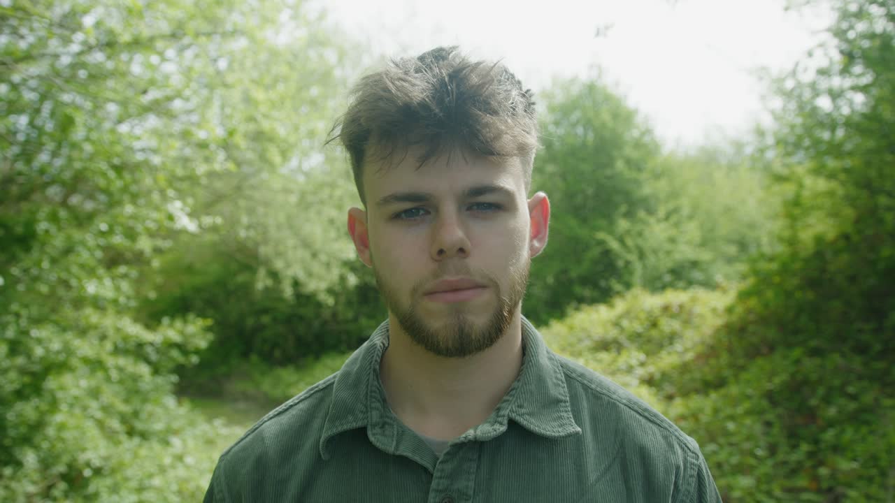 Close-up of man turning to side in forest clearing, framed by green leaves and afternoon light