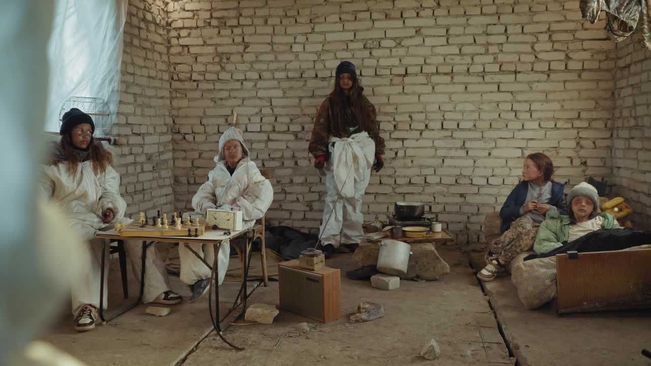 Stone age like survivors sit inside rundown shelter with cracked floor and tired faces, group tense as leader prepares to rise with cutlass, while makeshift kitchen and chessboard remain nearby