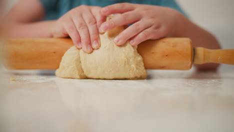 fair skin child rolling dough over wooden pin on floured white kitchen table during baking session, showing close up of small hands pressing dough with strength