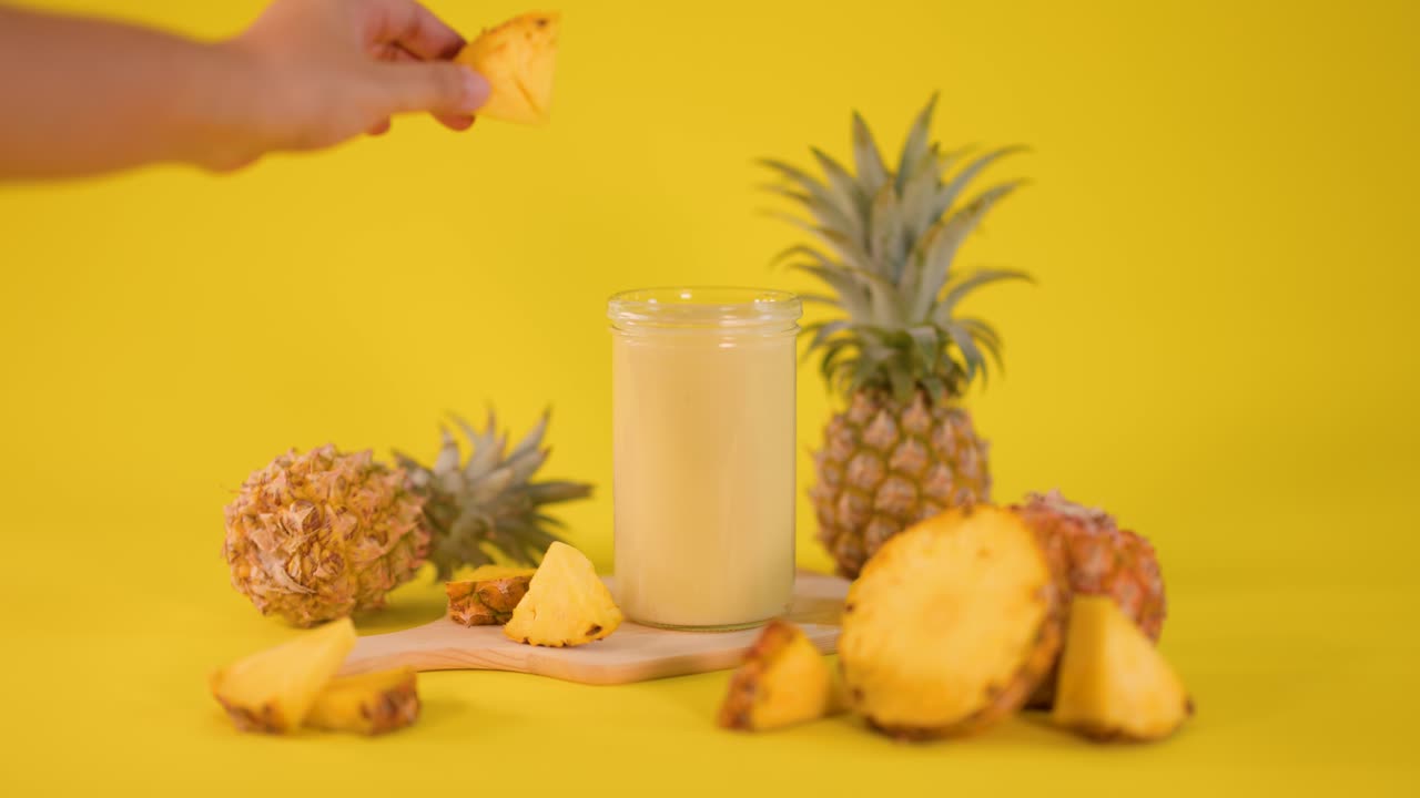 Hand places pineapple slice on creamy smoothie surrounded by fresh fruit, bright yellow studio lighting
