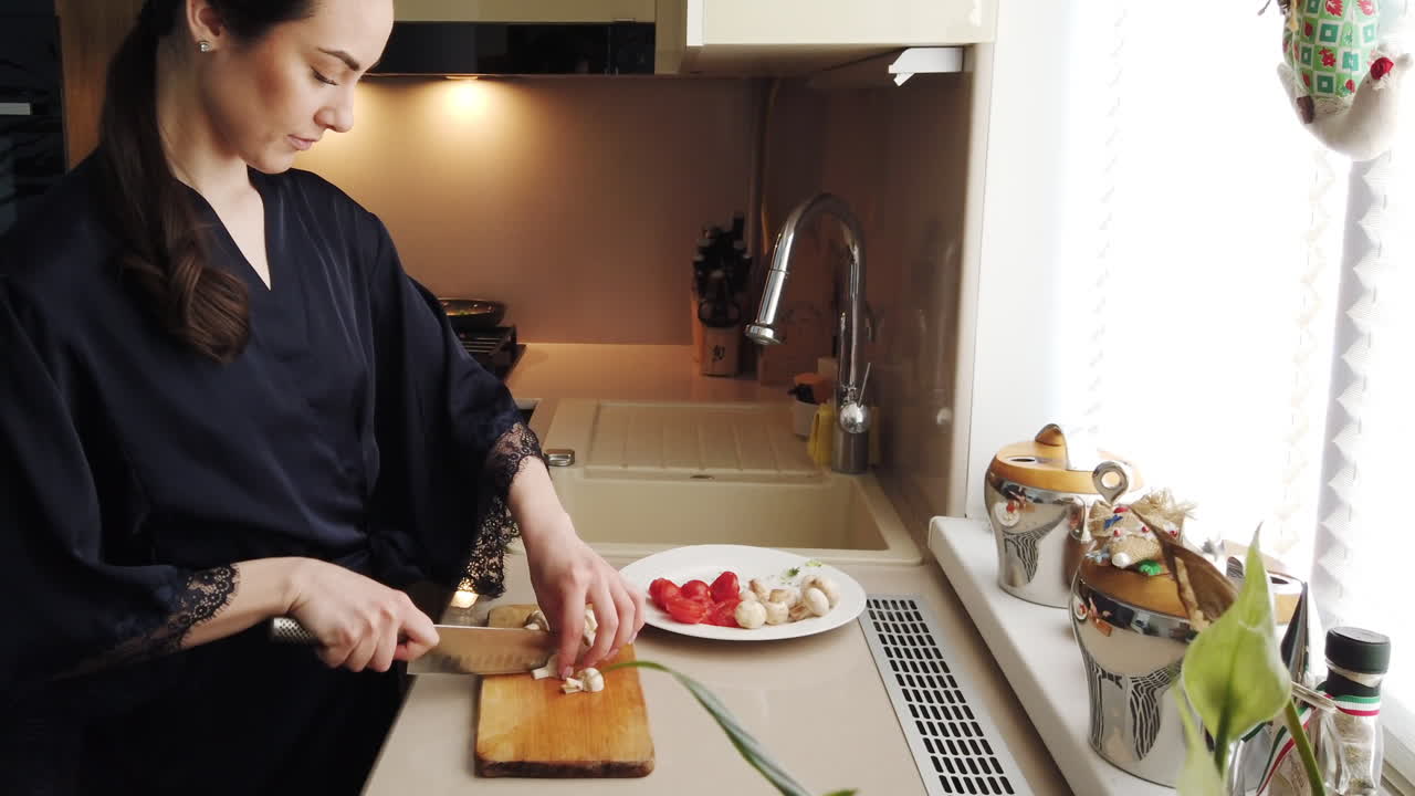 Woman in a blue robe cutting up mushrooms on a wooden board in the kitchen
