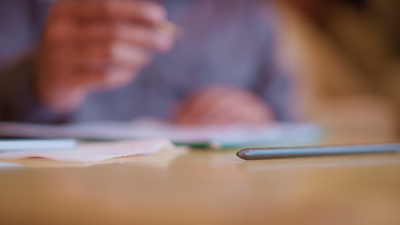 Blurred person in background writing on paper while pencil lies in sharp focus on wooden desk, subtle lighting enhances quiet workspace atmosphere, creating sense of concentration, stillness, and intention