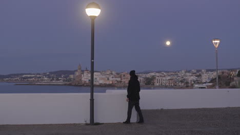 city-scape,-man-enters,-takes-photos-of-coastal-town,-as-moon-shines-above-and-light-switches-off