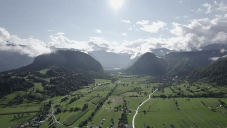 Frontales-Drohnenvideo,-Das-Sich-über-Farmen-Und-Häuser-Im-Triglav-Bewegt,-Mit-Hügeln-Am-Horizont,-Der-Sonne-Im-Vordergrund-Und-Einem-Klaren-Himmel-Mit-Wolken