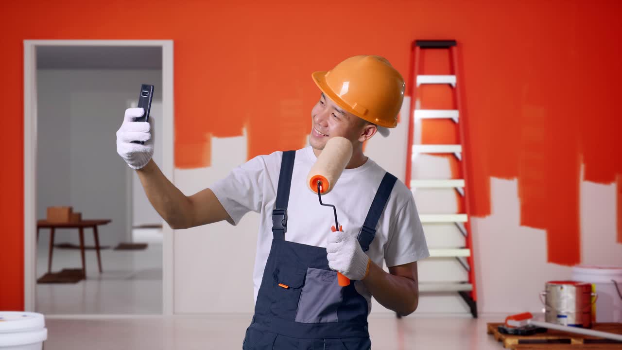 pintor asiático que usa un casco de seguridad usando un teléfono inteligente tomando una foto mientras está de pie con la pared de pintura en la habitación
