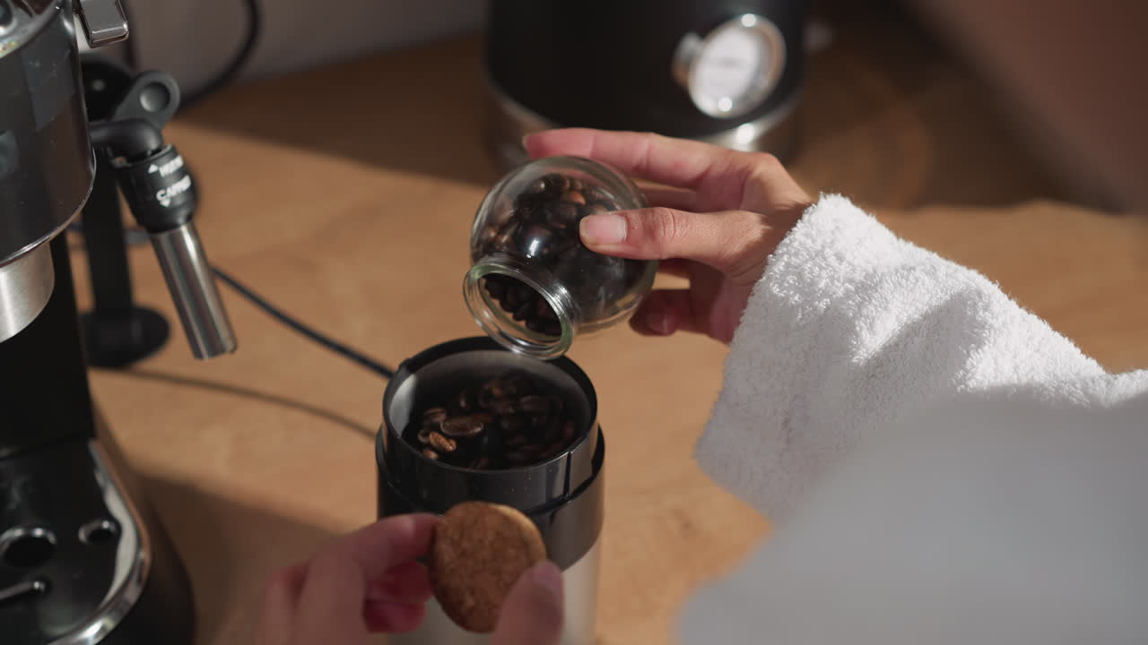 женщина добавляет кофейные бобы в мельницу за столом. дама кладет обжаренные зерна в кухонное устройство, начиная приготовление напитка. начинается ароматический ритуал