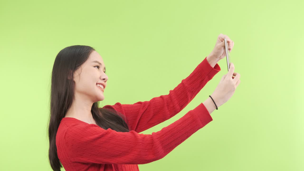 젊은 아시아 여성이 휴대전화를 사용하여 녹색 화면 배경으로 셀피를 찍습니다.