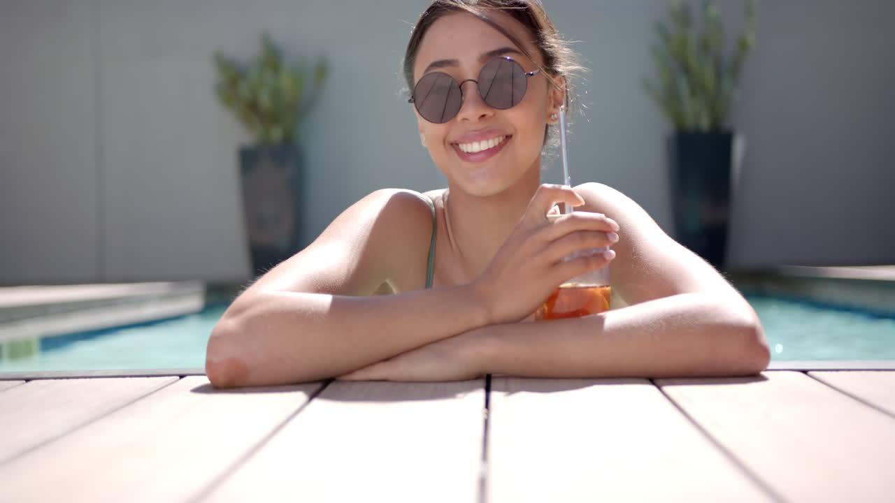 Smiling woman in sunglasses relaxing by pool, holding refreshing drink