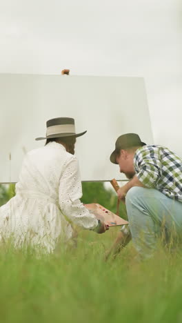 two individuals bent over in a grassy field, focused on their tasks. the woman in a white dress holds a palette, while the man in a checked shirt and hat reaches for something on the ground