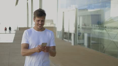 Smiling-bearded-guy-walking-with-smartphone-near-modern-building