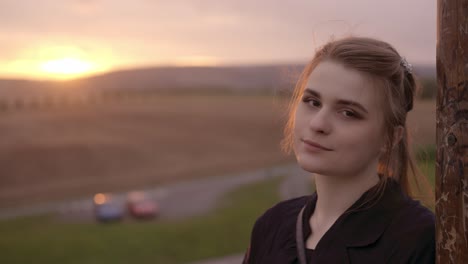 smiling girl with brown eyes and hair leaning on a pillar with a romantic summer sunset background