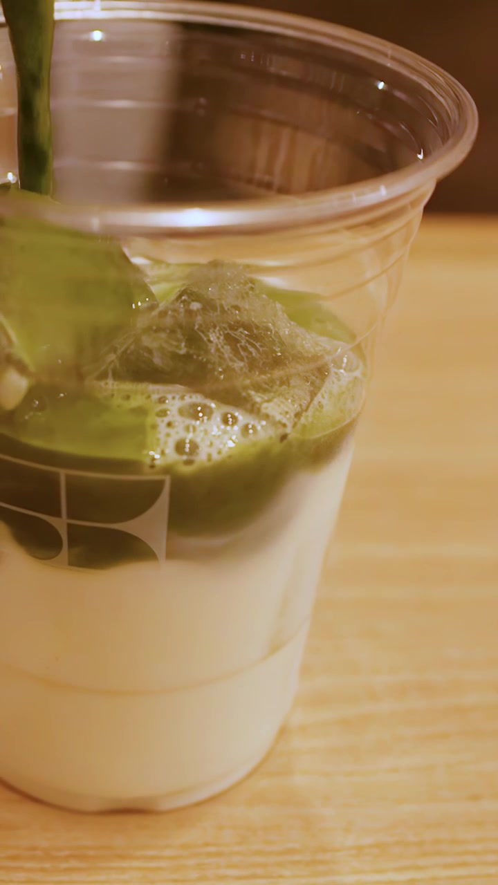 Tiivistetty vihreä matcha-tee valuu muovikuppiin, joka on täynnä maitoa ja jääpaloja.