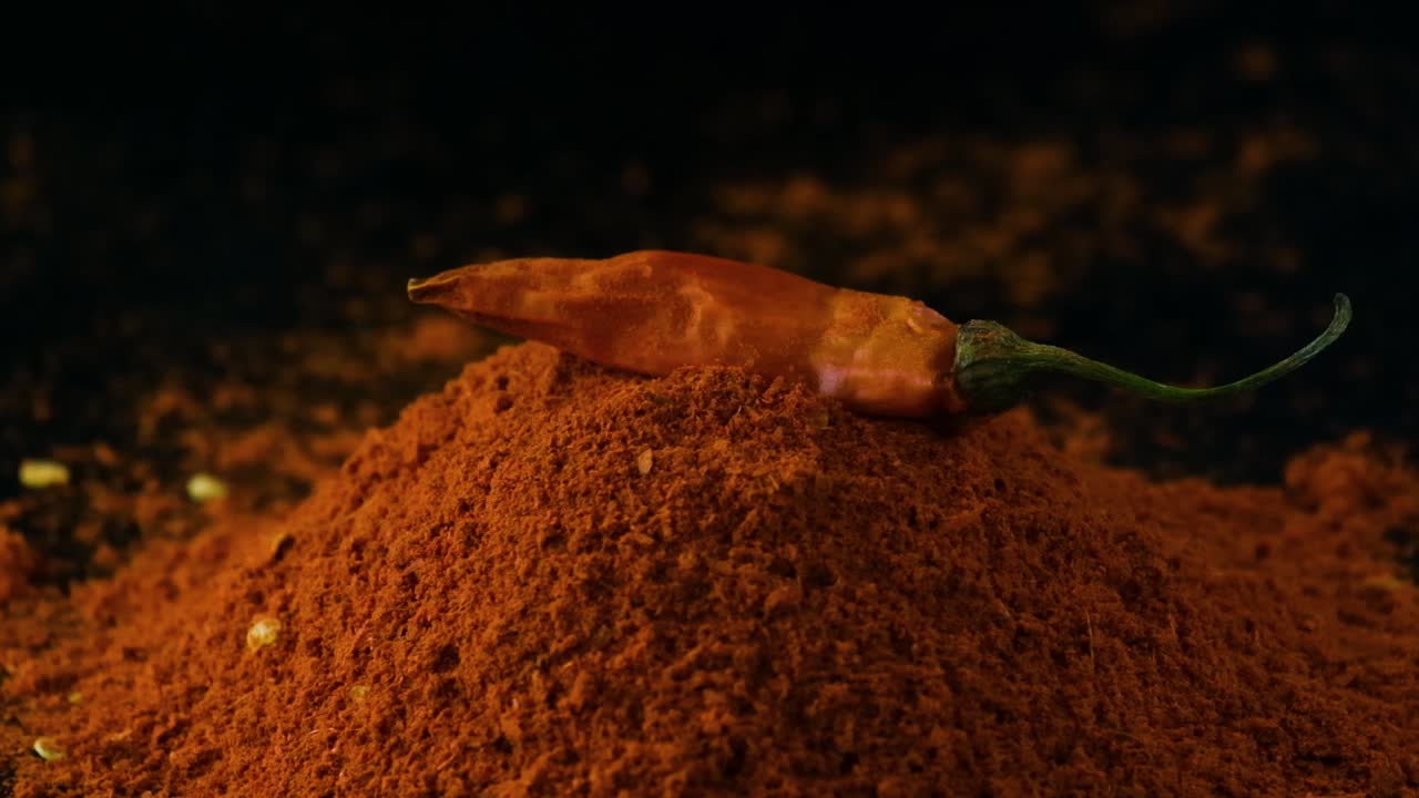 Slow motion whole red chilly pepper falling onto pile of savoury paprika powder isolated on black close up